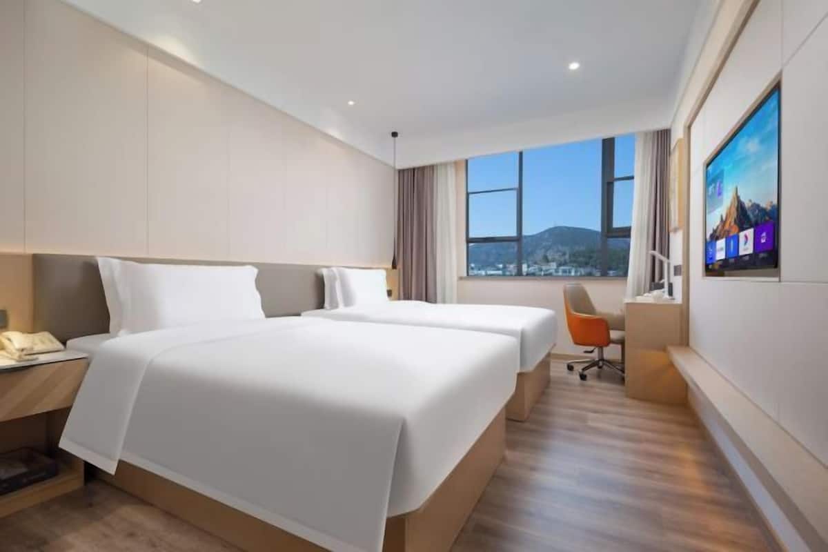 A hotel room with two beds, a desk, a chair, a TV, and a view of the city.