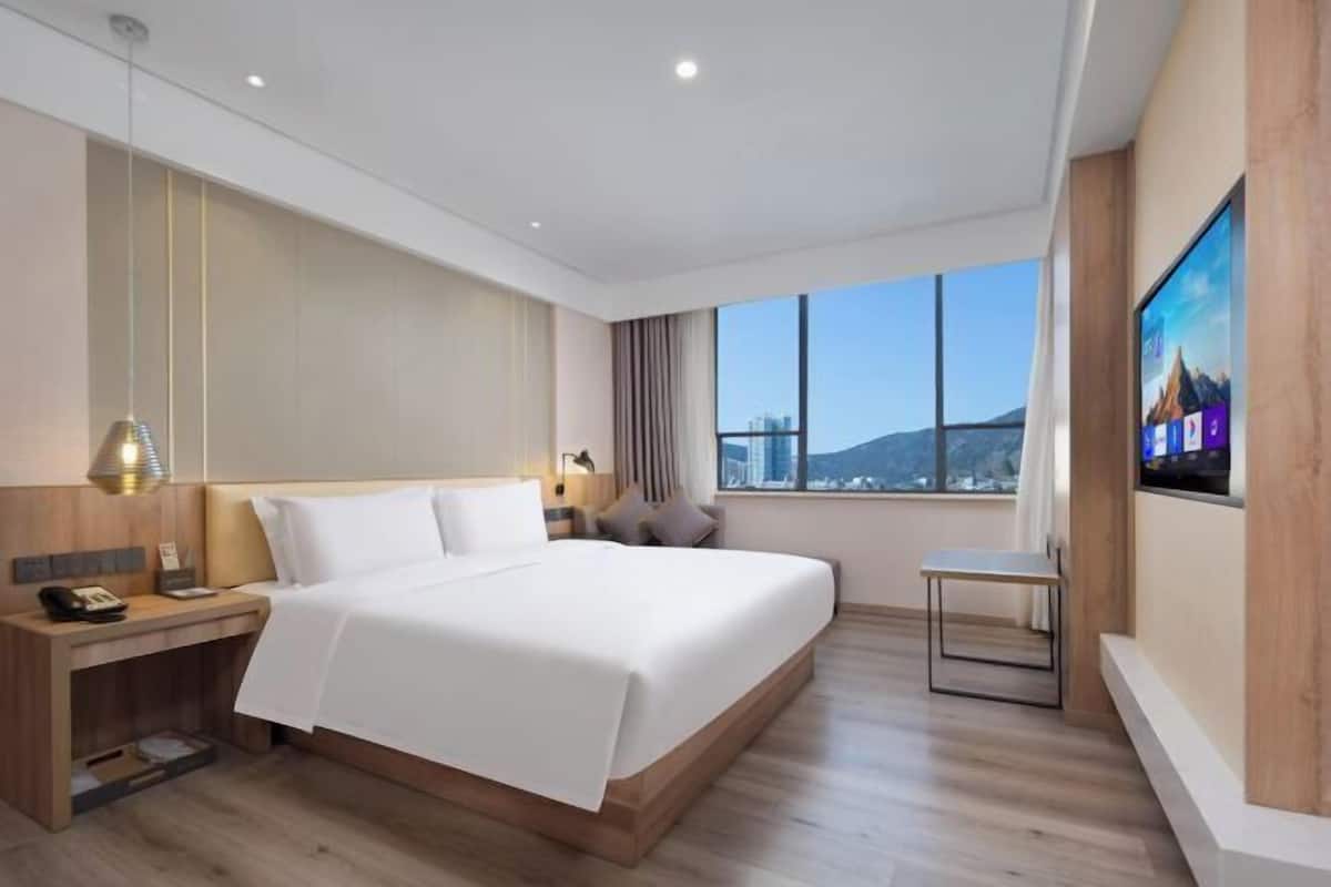 A modern hotel room with a large bed, a desk, a chair, a TV, and a city view.