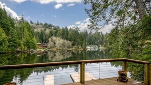 Una terraza de madera con barandilla da vista a un lago tranquilo, rodeado de un bosque denso y algunos edificios a lo lejos.
