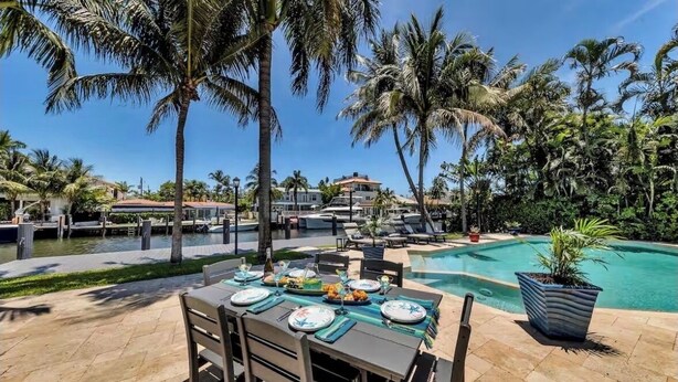 A poolside dining area with a table set for a meal, surrounded by palm trees and a clear blue sky.