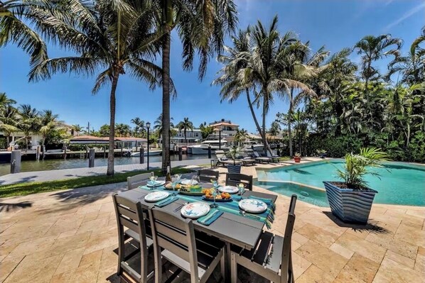 Un área de comedor junto a la piscina con una mesa dispuesta para una comida, rodeada de palmeras y un cielo azul despejado.