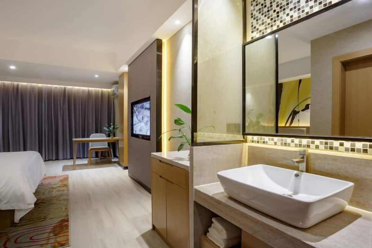 A modern hotel room with a bed, a desk, a television, a bathroom with a large mirror and a sink.