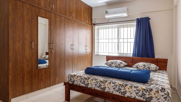 A bedroom with a wooden wardrobe, a bed with floral bedding, a window with curtains, and an air conditioning unit. - Pinnacle Serviced Apartments (Bengaluru)