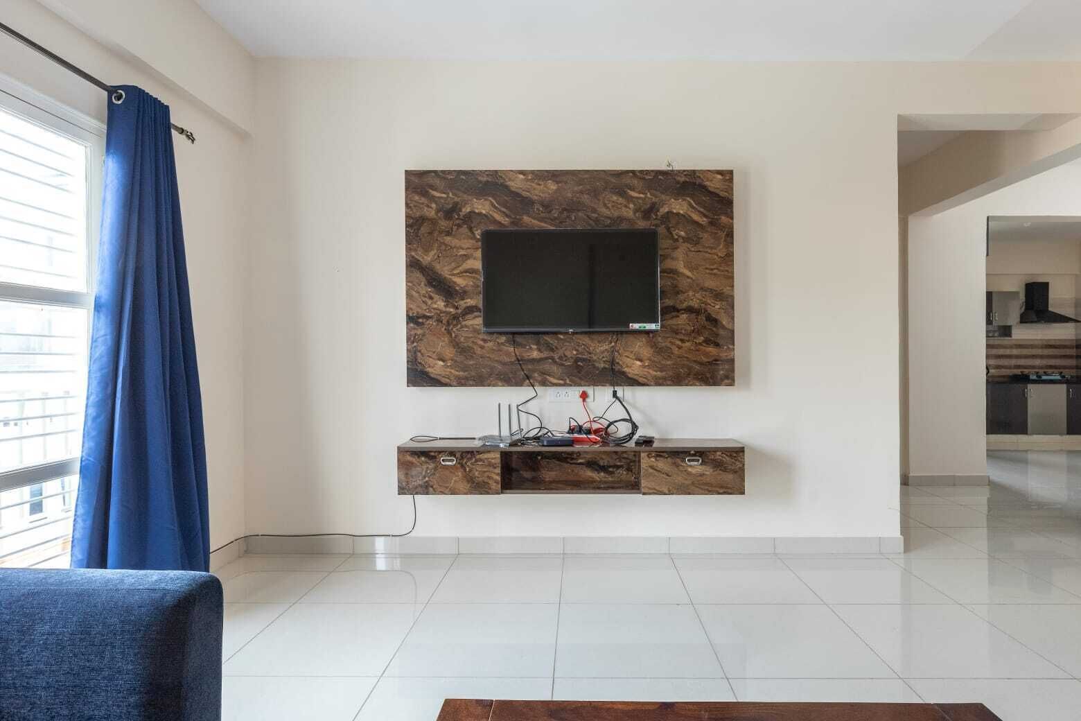 A modern living room with a flat-screen TV mounted on a marble-patterned wall, a wooden entertainment unit below, and a blue curtain on the window.