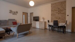 Ein modernes Wohnzimmer mit einer Couch, einem außergewöhnlichen Sessel, einer Essecke mit Tisch und Stühlen, einem Fernseher auf einer Holzkommode und einer Wand mit Steinakzenten.