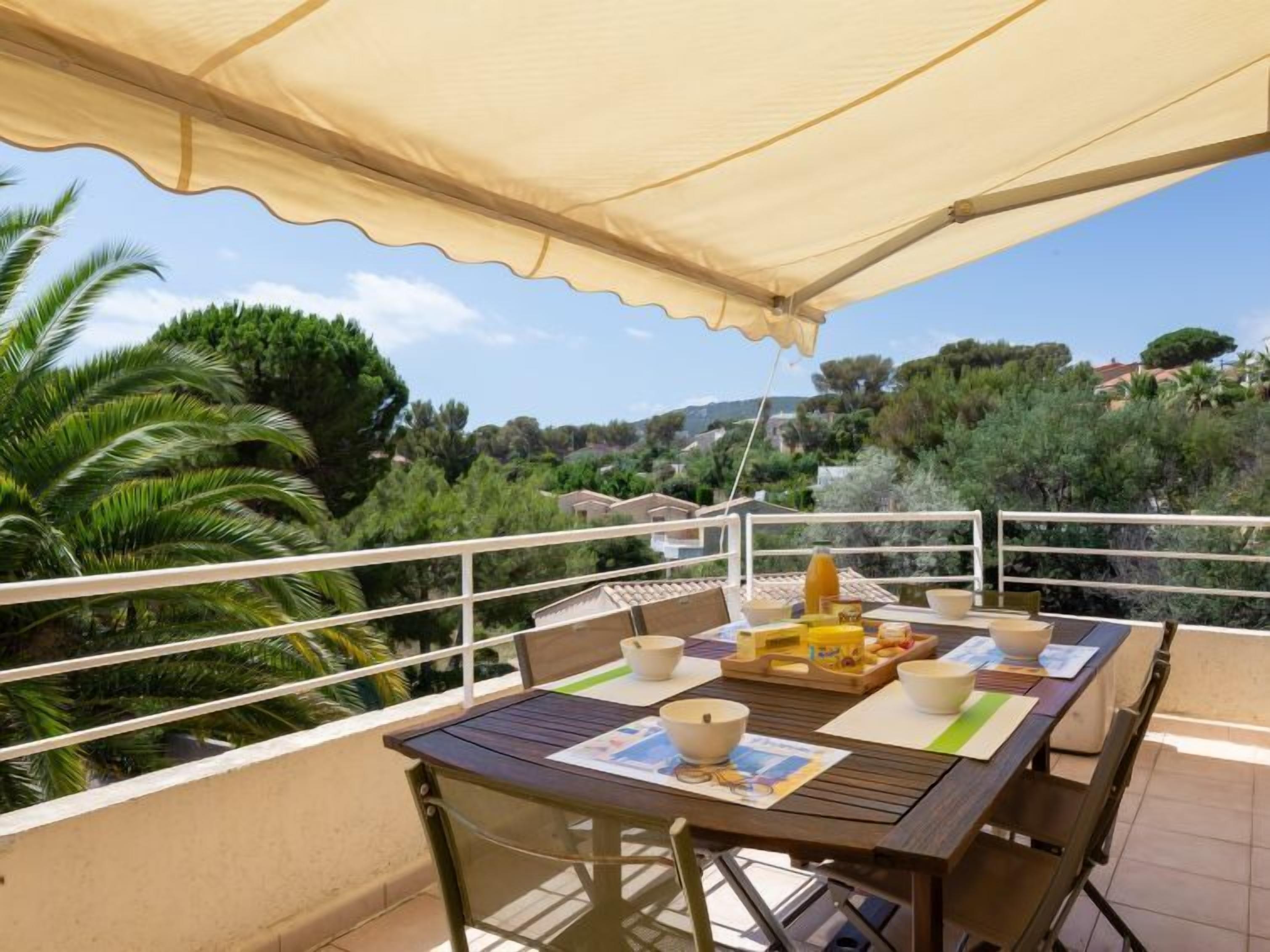 Un balcon avec une table dressée pour le petit-déjeuner, entouré d’une balustrade blanche, donnant sur un paysage luxuriant parsemé d’arbres et de maisons.
