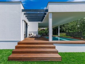 Ein moderner Poolbereich im Freien mit einer Holzterrasse, die zu einer Sitzgruppe unter einer Pergola führt.