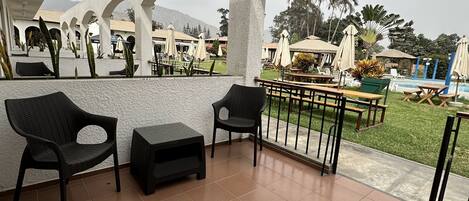 Un espacio de estar al aire libre techado, con sillas y mesas, con vistas a una piscina y vegetación.