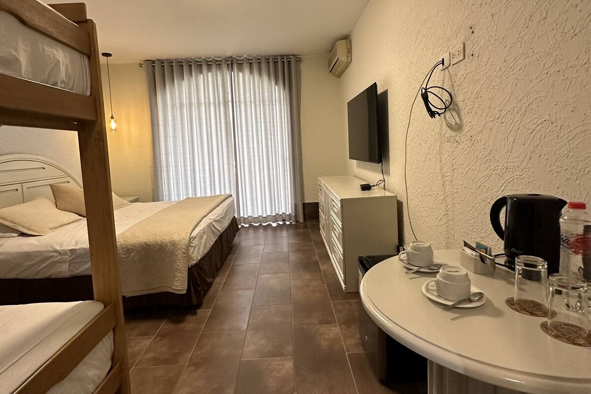 Una habitación de hotel con dos literas, un escritorio con un televisor de pantalla plana, un aparador y una mesa redonda con una cafetera y tazas.