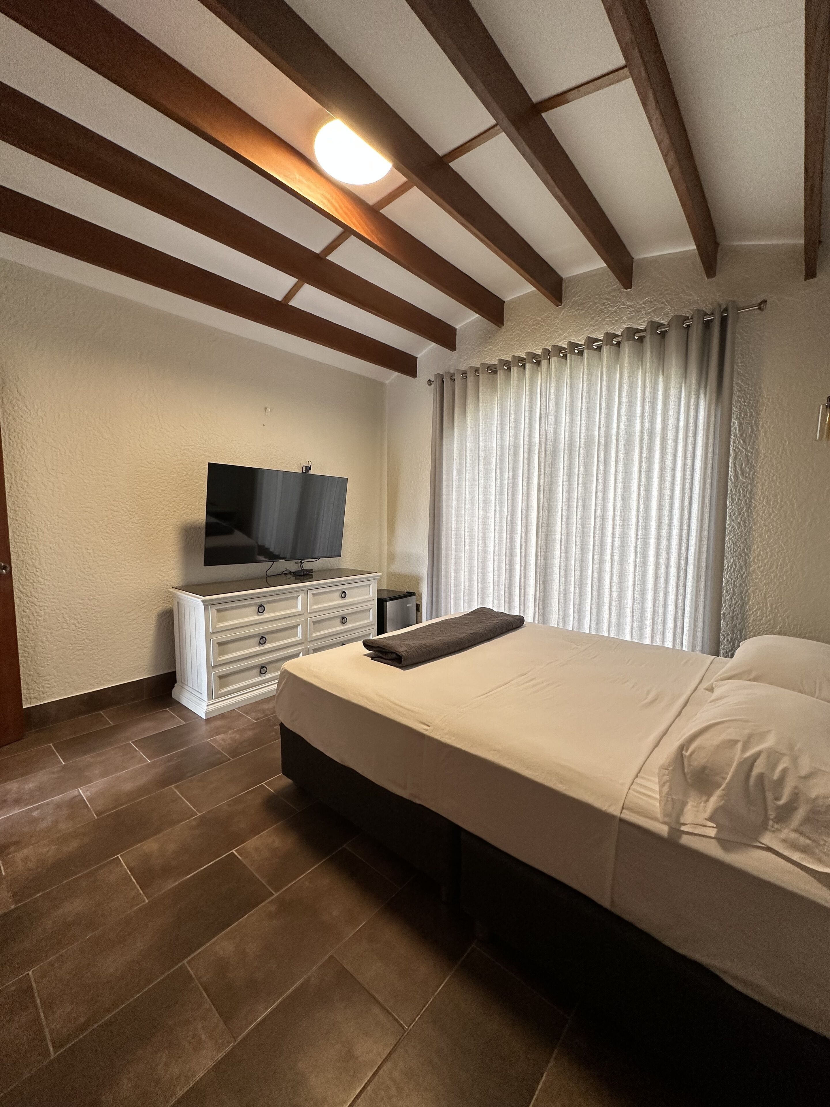 A bedroom with a bed, a TV on a dresser, and a window with curtains.