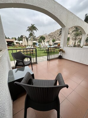 A covered outdoor seating area with wicker chairs and tables, a view of a mountainous landscape, and a patio with umbrellas.