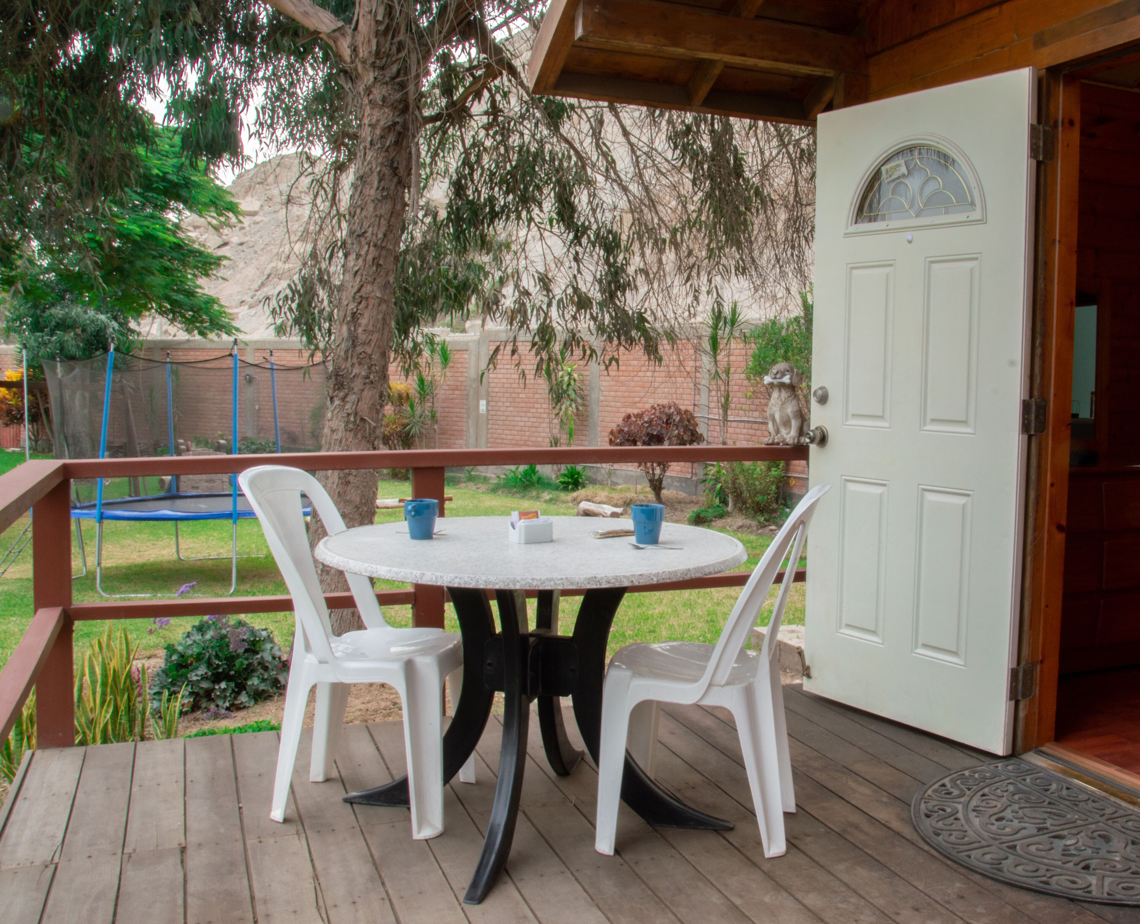 A wooden deck with a round table and four white chairs, a white door with a glass window, and a small dog statue on the door.