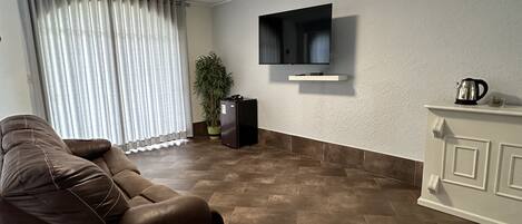 A living room with a brown leather sofa, a small white table, a flat-screen TV mounted on the wall, a potted plant, and a cabinet with a kettle on top.