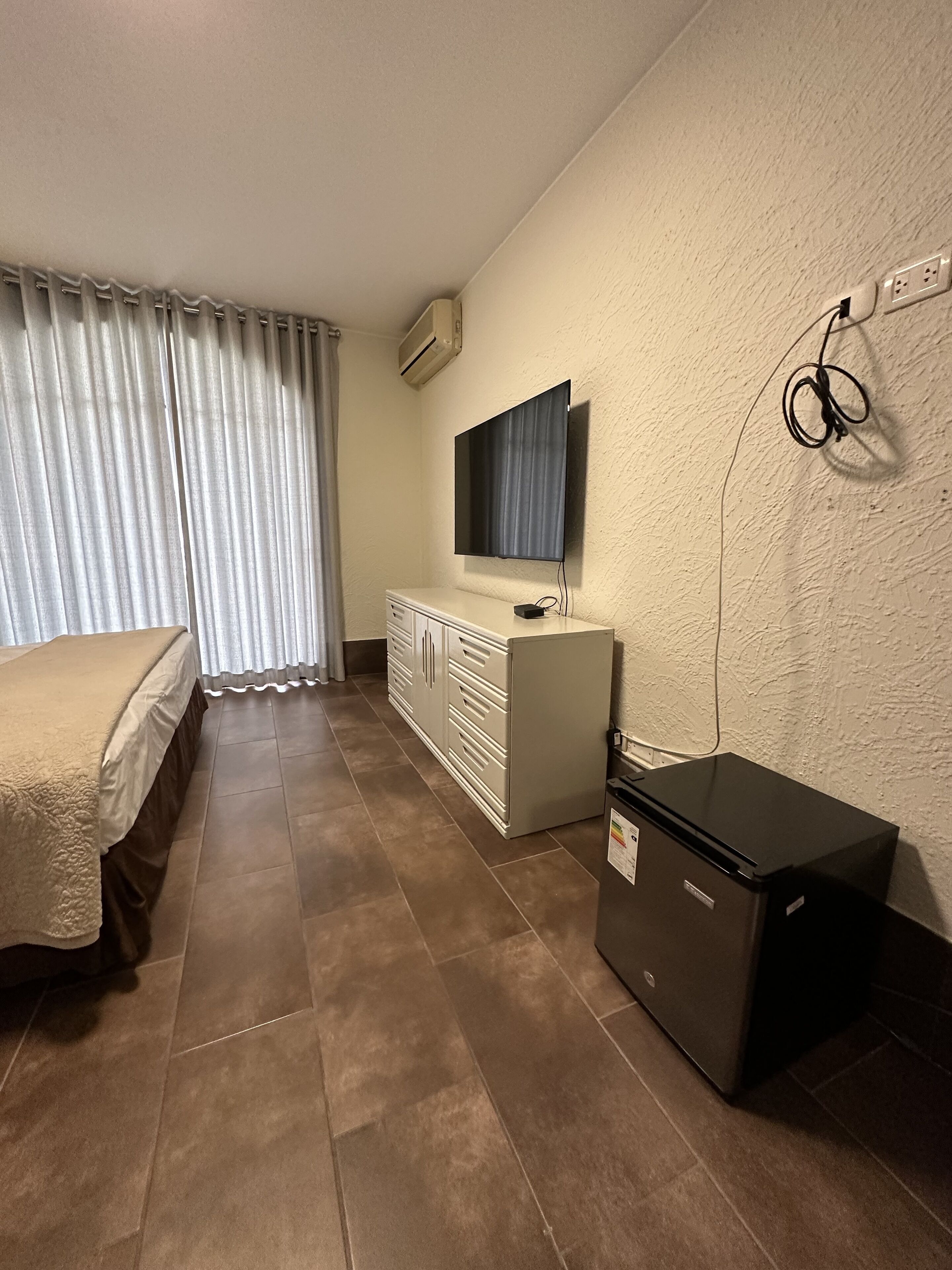 Una habitación de hotel con una cama, un aparador, un televisor montado en la pared y un refrigerador pequeño.