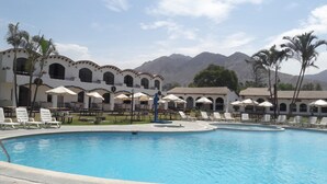 Zona de piscina con sillones de descanso y sombrillas, un edificio con arcos y montañas al fondo.