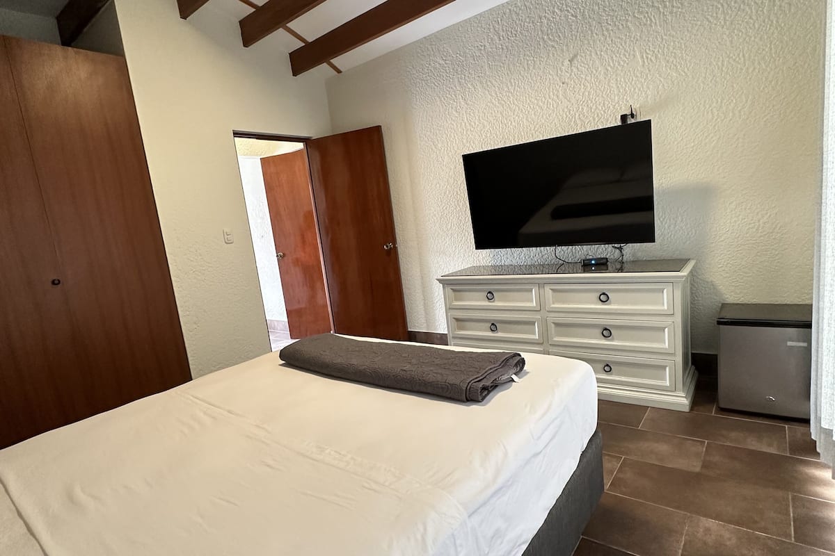 Un dormitorio con cama, televisor sobre una cómoda, una puerta y piso de baldosas.
