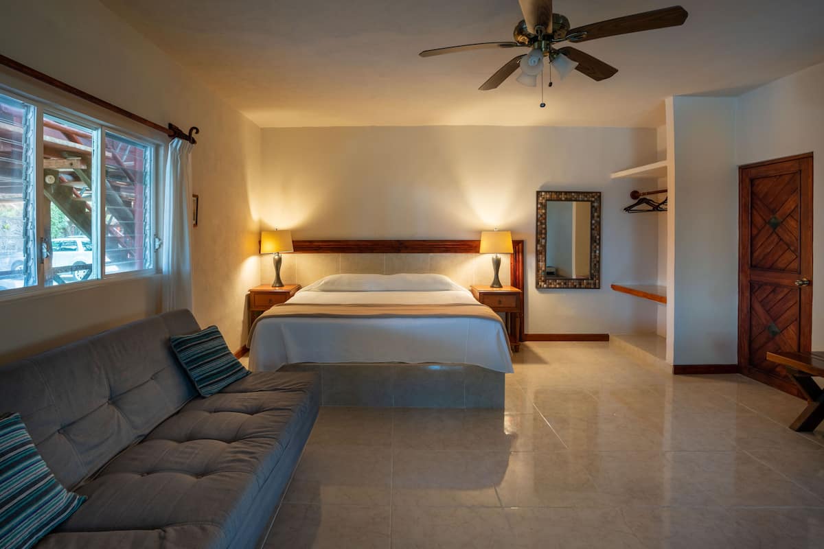 A bedroom with a bed, a sofa, a ceiling fan, a mirror, and a door.