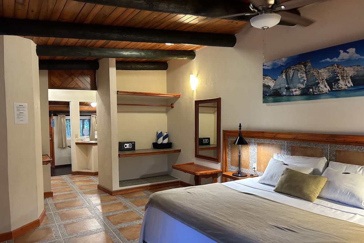 A hotel room with a bed, a nightstand, a lamp, a mirror, and a painting of a mountain landscape.