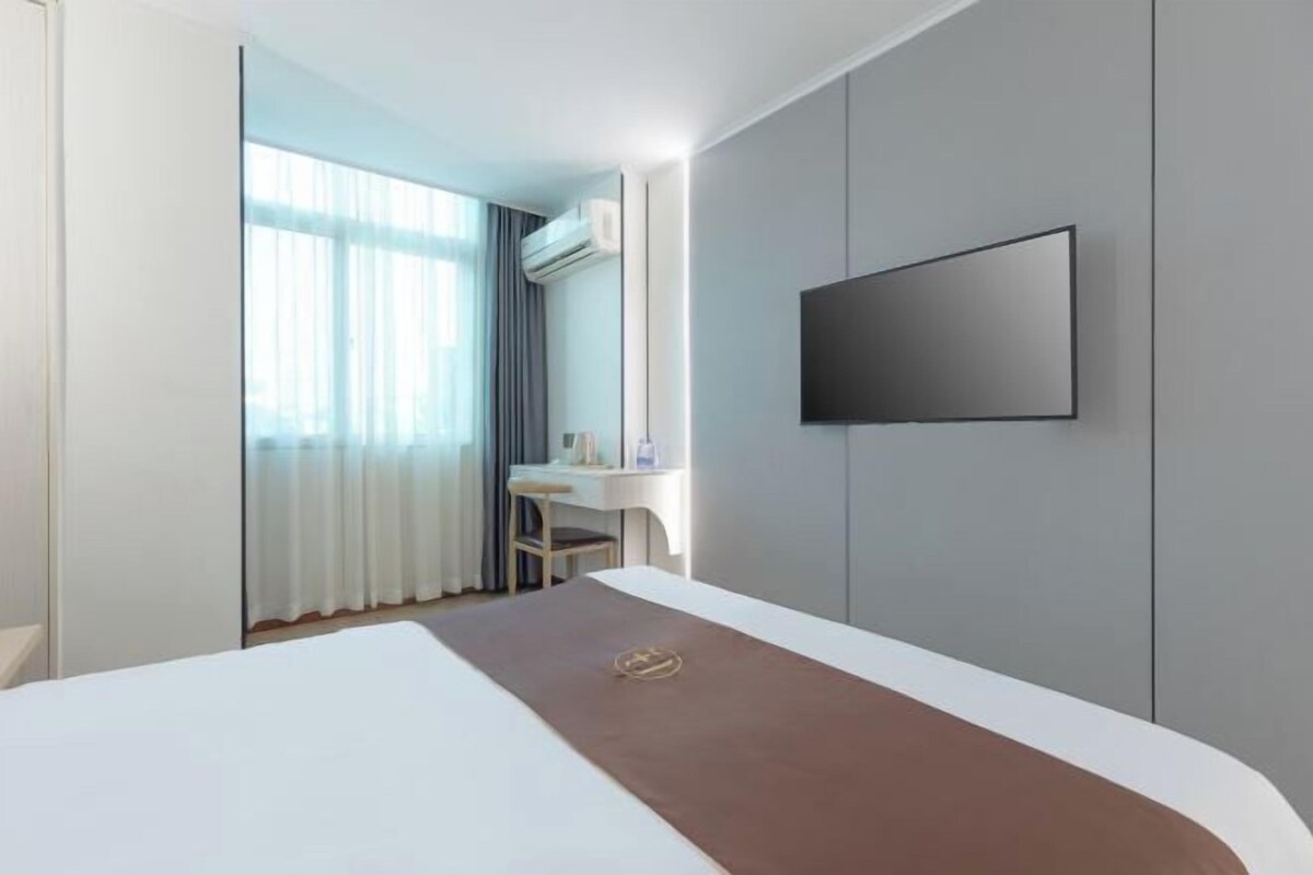 Ett hotellrum med en säng, ett skrivbord, en stol, en tv som är monterad på väggen och ett fönster med gardiner.