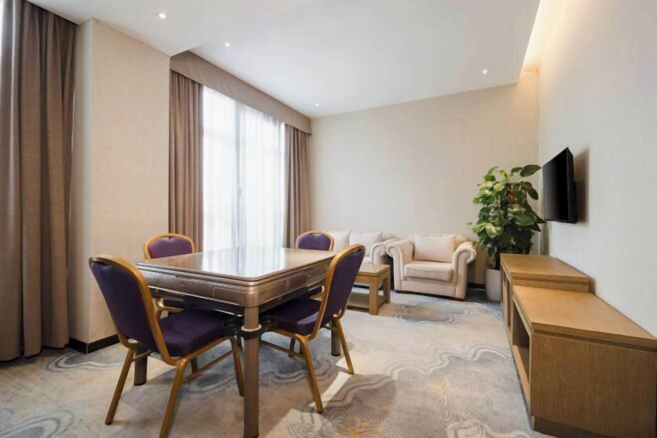 A hotel room with a dining table, two chairs, a sofa, a TV mounted on the wall, and a plant.