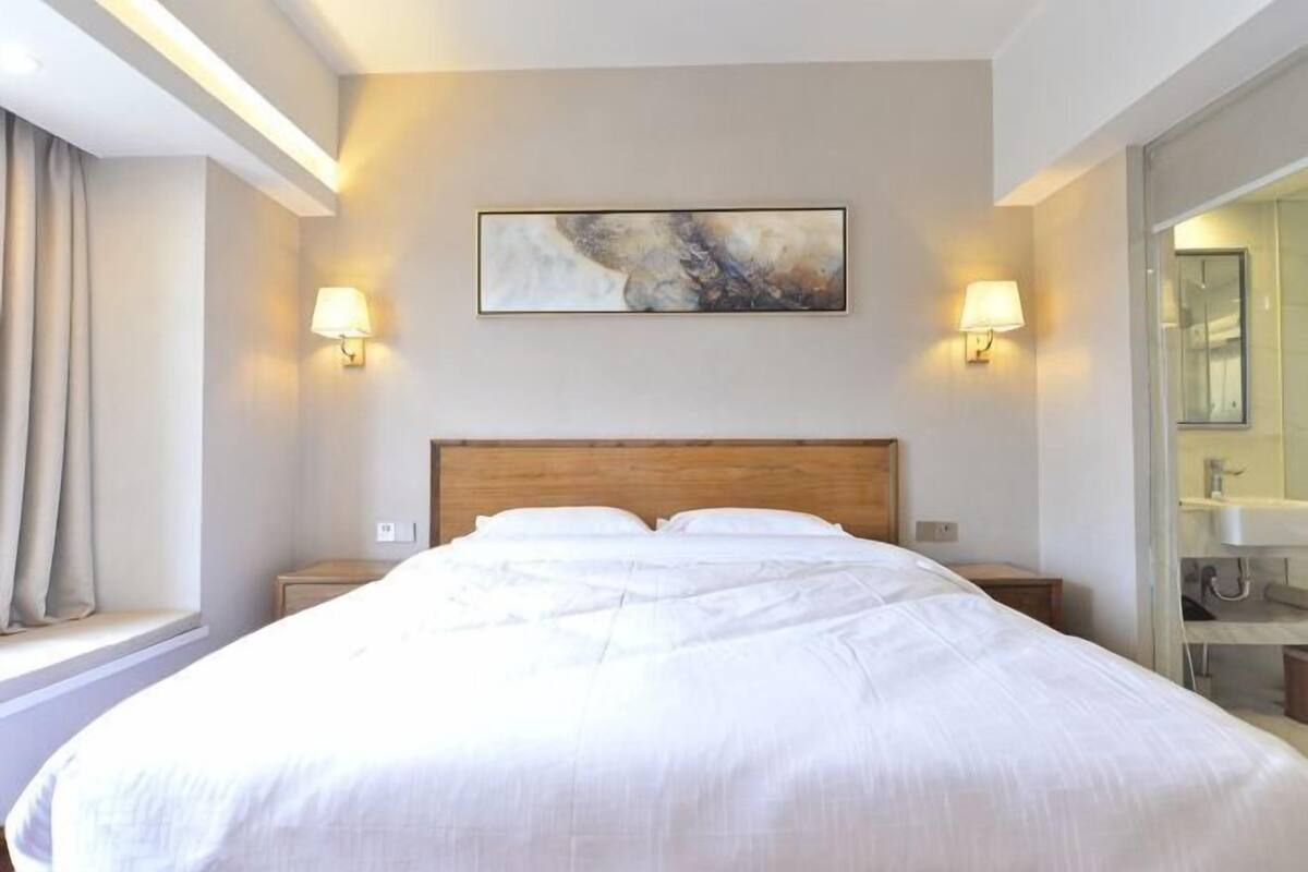 A hotel room with a large bed, two bedside lamps, a painting on the wall, and a bathroom visible through an open door.