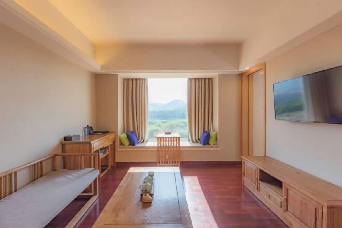 A room with a wooden bench, a small table with a teapot, a TV on the wall, and a window with curtains.