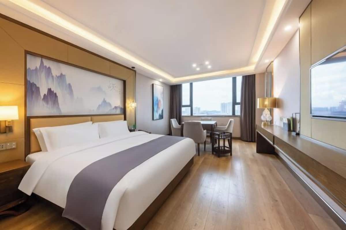 A hotel room with a large bed, a desk with a chair, a window with a city view, and a painting on the wall.