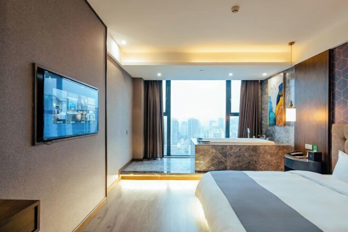 Moderni hotellihuone, jossa on suuri sänky, seinälle asennettu televisio, kylpyammeella varustettu kylpyhuone ja näkymä kaupunkiin.