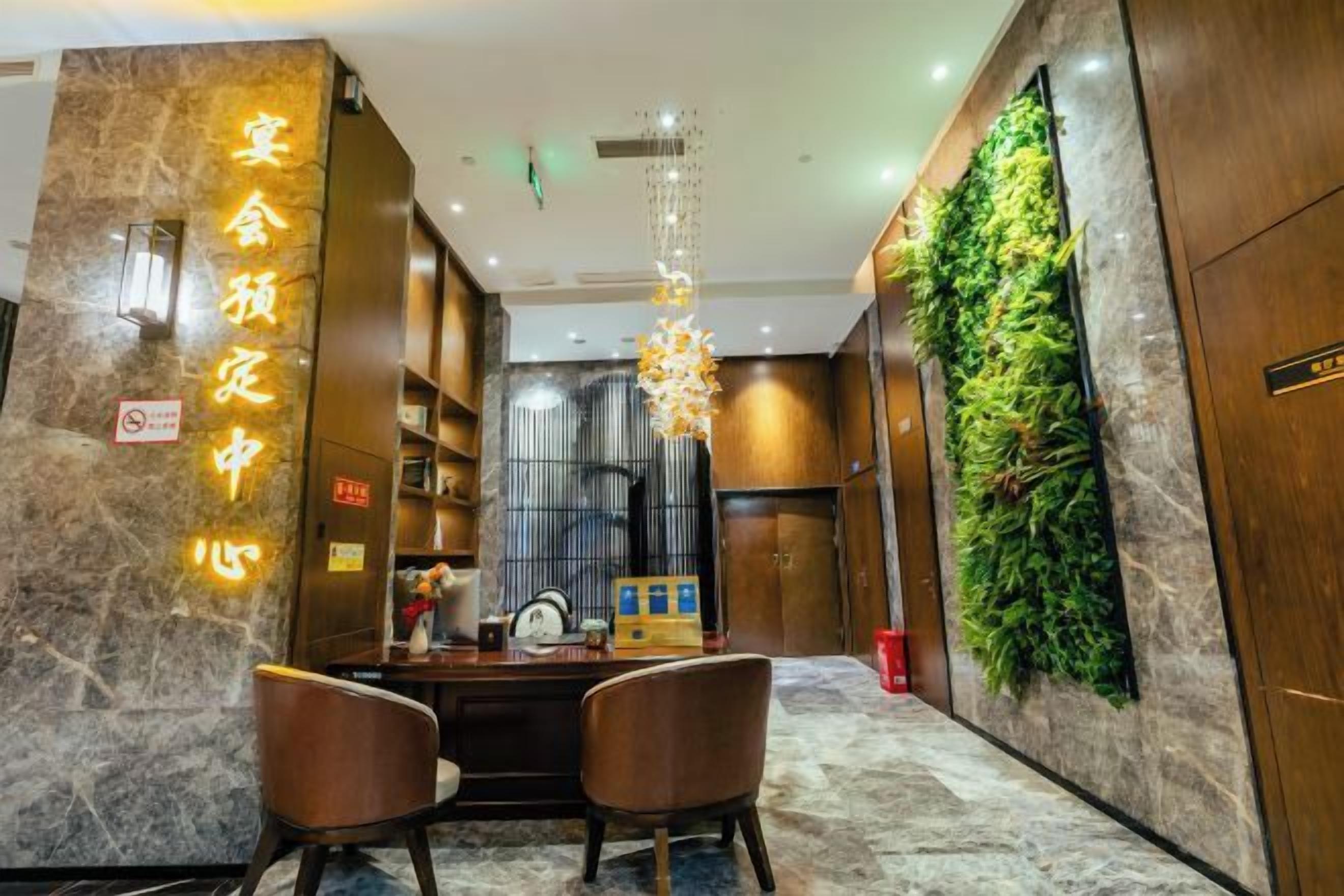 A hotel lobby with a reception desk, two chairs, a chandelier, a wall-mounted plant, and a marble floor.