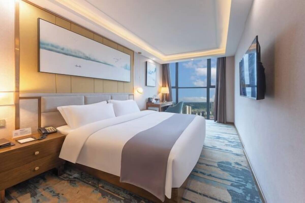 A hotel room with a large bed, a nightstand, a desk, a chair, a TV mounted on the wall, and a large window offering a city view.