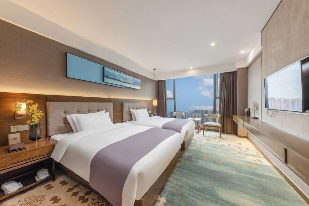 A hotel room with two beds, a large TV, a city view, and a desk with a lamp.