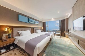 A hotel room with two beds, a large TV, a city view, and a desk with a lamp.
