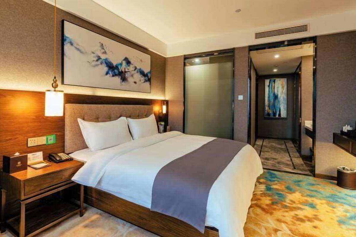 A hotel room with a bed, bedside table, lamp, artwork, and a view of the hallway.