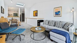 Un soggiorno moderno con un divano grigio, poltrone blu e un tavolino da caffè. Si distingue una zona pranzo con un tavolo e sedie in legno, e una cucina sullo sfondo.