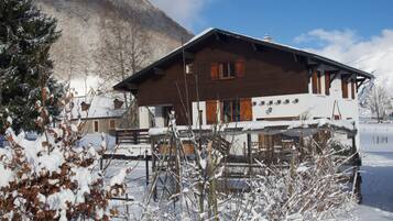 A wooden chalet with a balcony, surrounded by snow-covered trees and a mountain in the background.
