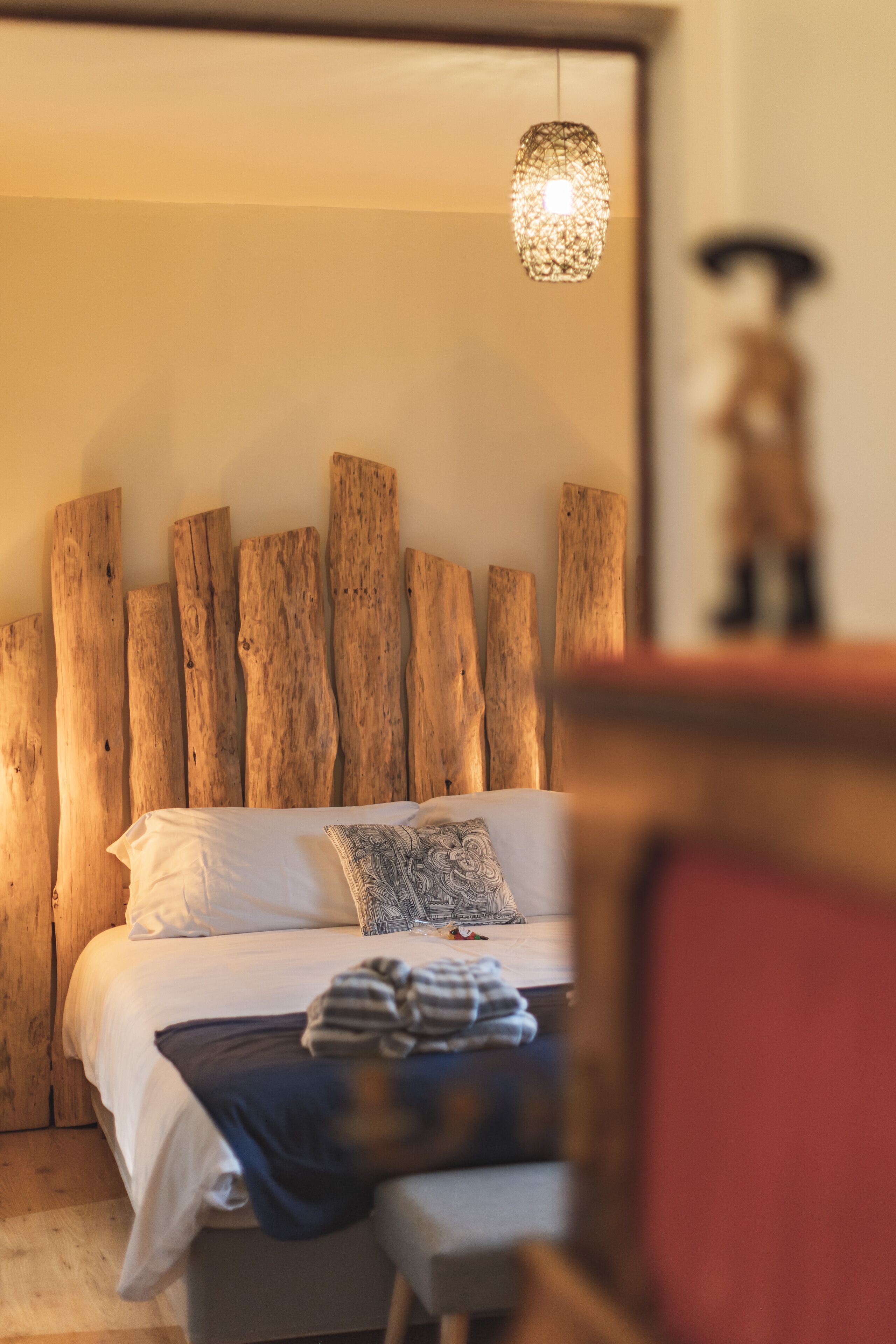 Ein Schlafzimmer mit einem hölzernen Kopfteil, einer Pendelleuchte und einem Bett mit weißer Bettwäsche und einem gemusterten Kopfkissen.