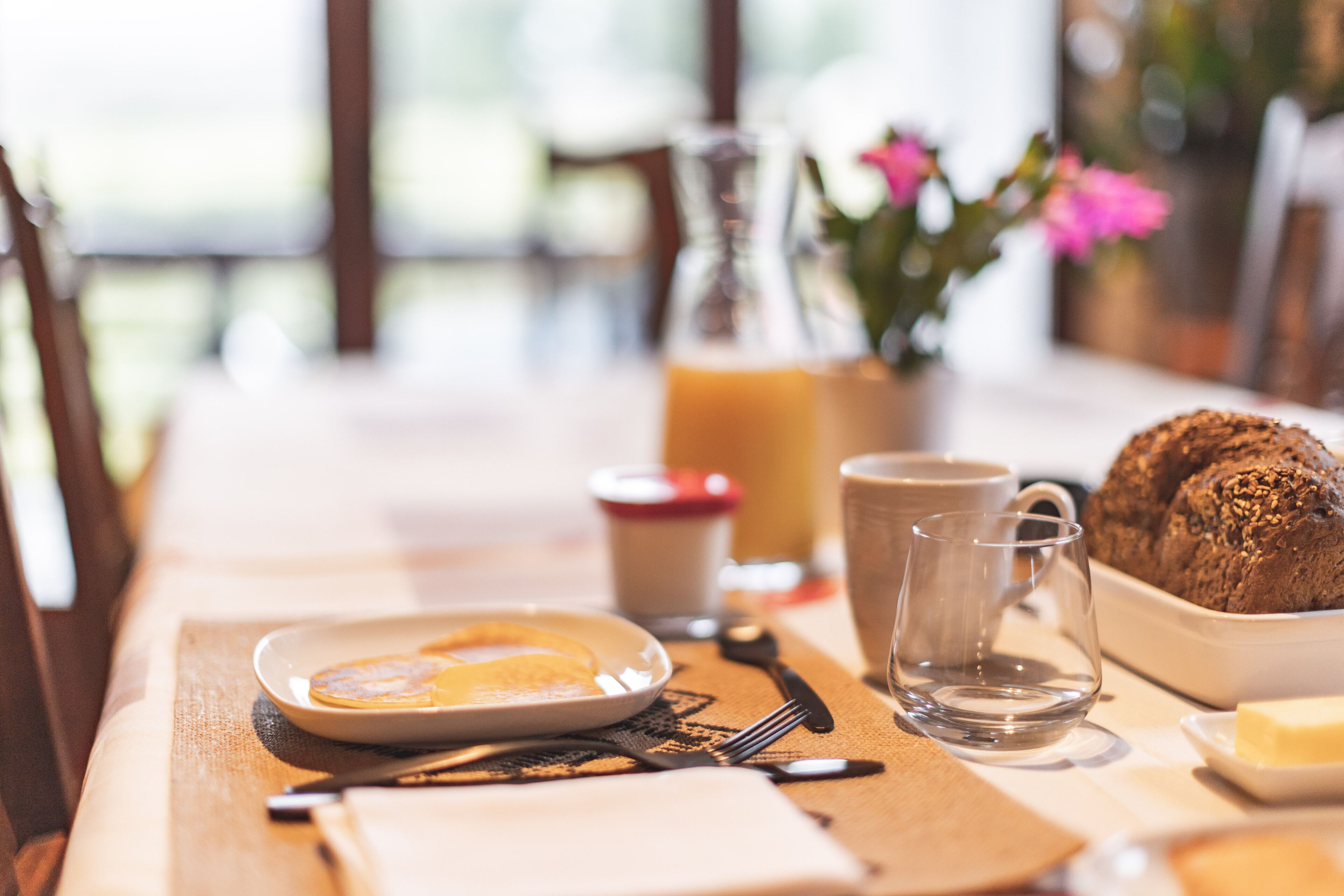 Une table de petit-déjeuner avec une assiette de nourriture, un verre de jus, une tasse, une bouteille de jus d’orange, un bol de muffins et une petite assiette avec du beurre.