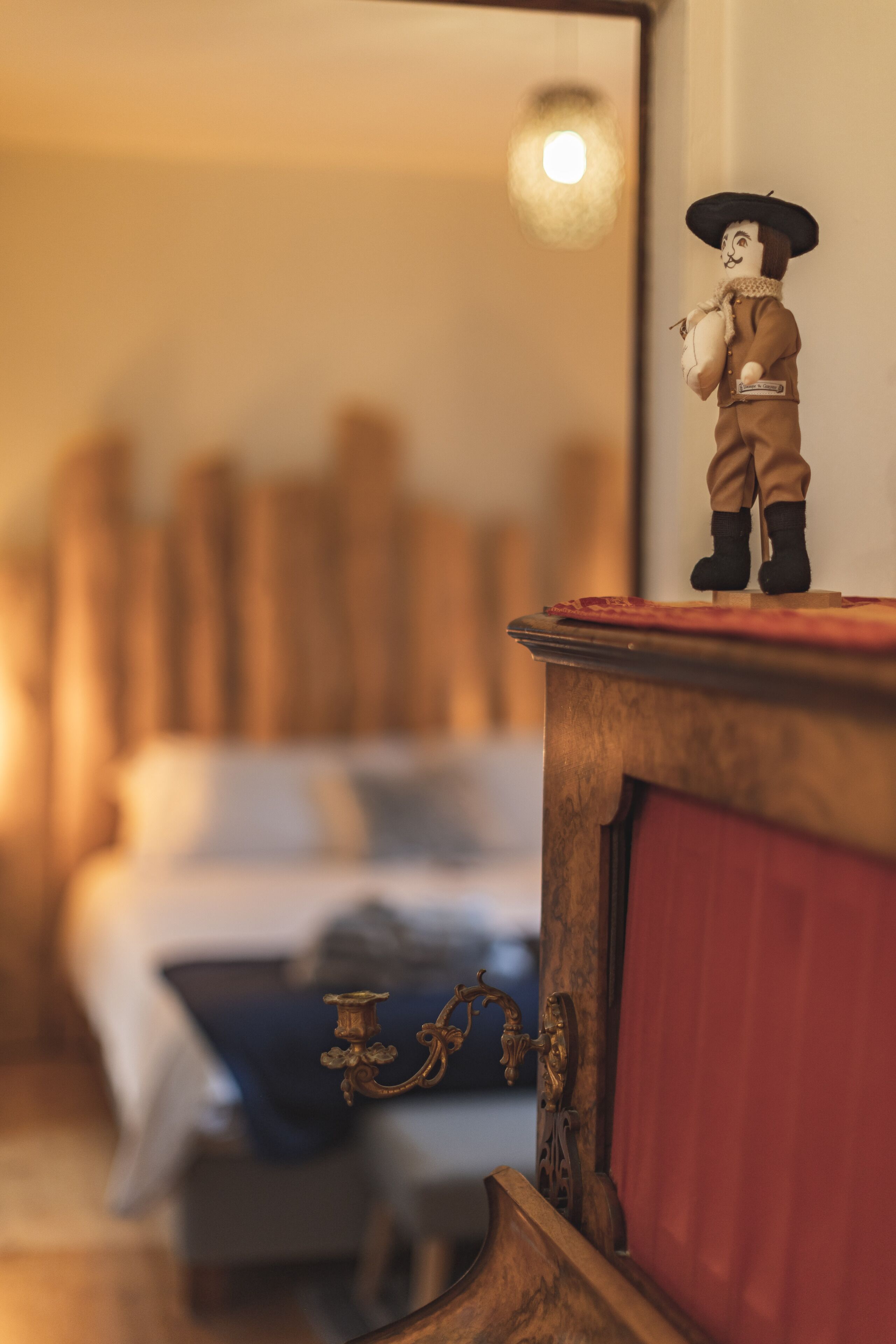 Een bed met een houten hoofdeinde, een nachtkastje met een beeldje, en een rood gordijn.