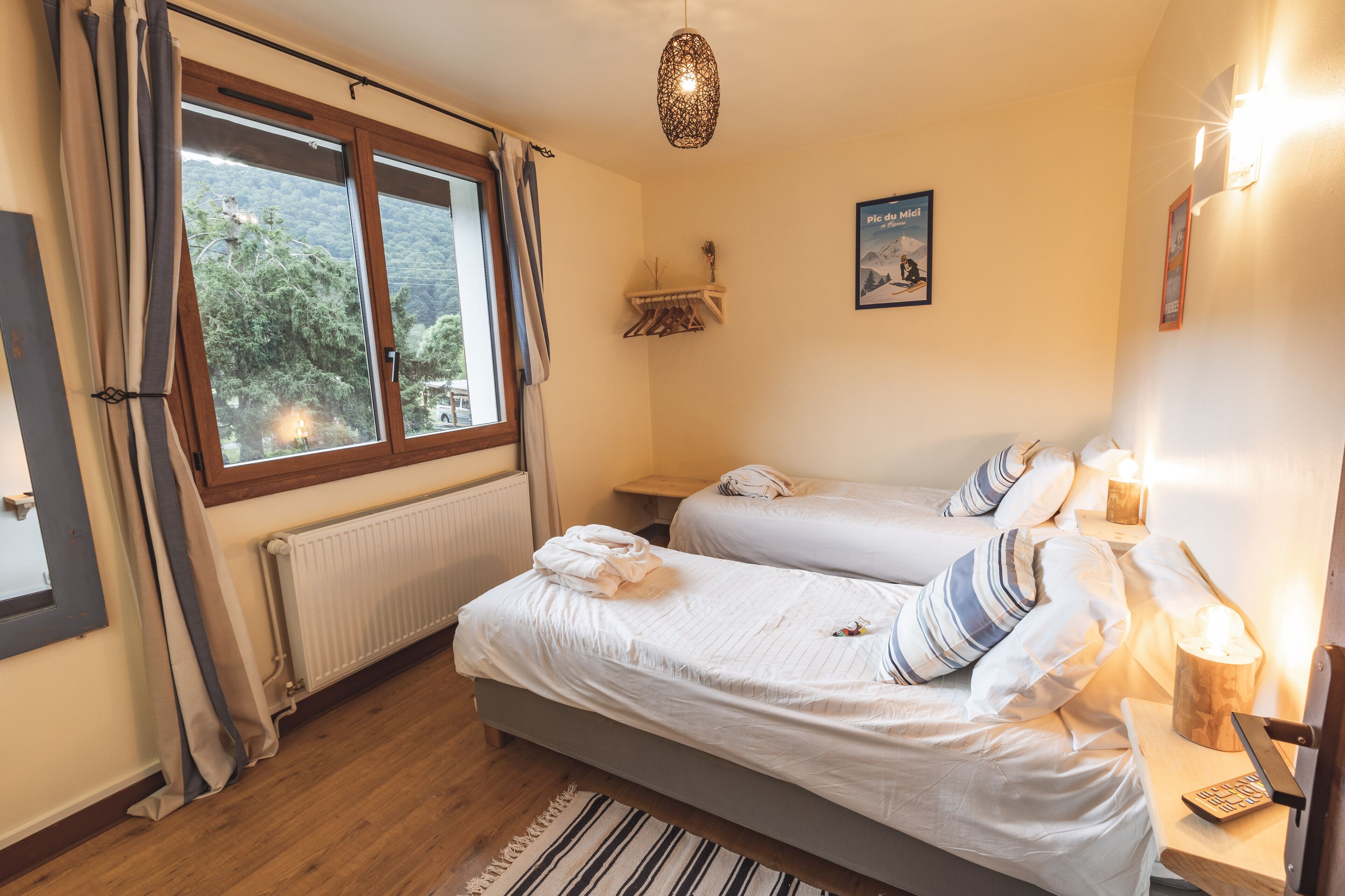 Ein Schlafzimmer mit zwei Betten, einem Fenster mit Vorhängen, einem Heizkörper, einem Nachttisch mit Lampe und einem an der Wand befestigten Regal mit einem Bild.