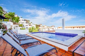 Un área de piscina en la azotea con sillas de descanso y un jacuzzi, rodeada de edificios y un cielo despejado.