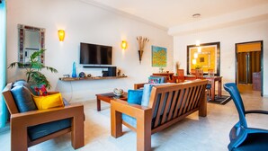 Un salon moderne avec un canapé, des fauteuils, une table basse et un téléviseur. Il y a également un coin repas avec une table et des chaises. La pièce est équipée d’un téléviseur à écran plat fixé au mur, d’une étagère avec des objets décoratifs et d’un tableau.