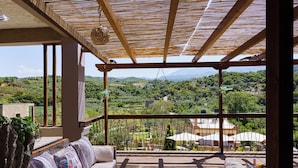A balcony with a wooden floor, a cushioned sofa, and a hanging chair. A view of a green landscape with hills and buildings is visible through the balcony. - Villa Fisi 1 - Pool, Hot Tub & Mountain View (Mylopotamos)