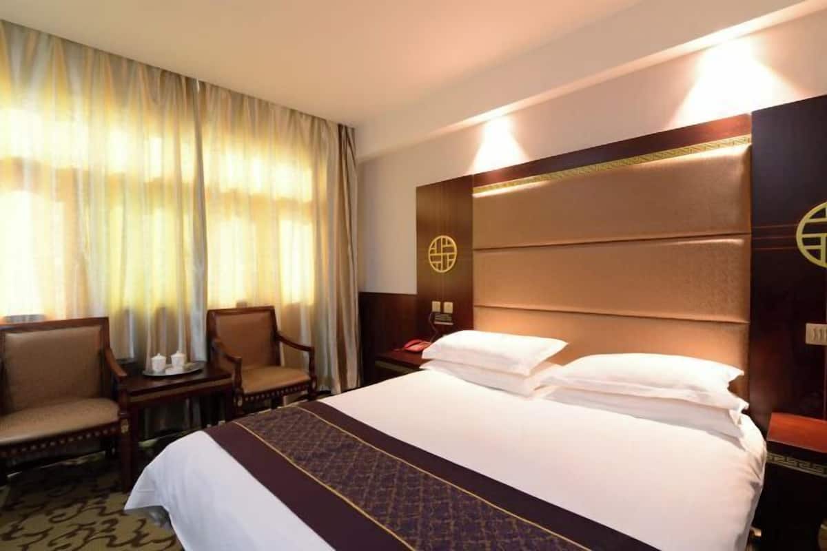 Ein Hotelzimmer mit einem Bett, zwei Stühlen, einem Nachttisch und einem großen Fenster mit Vorhängen.