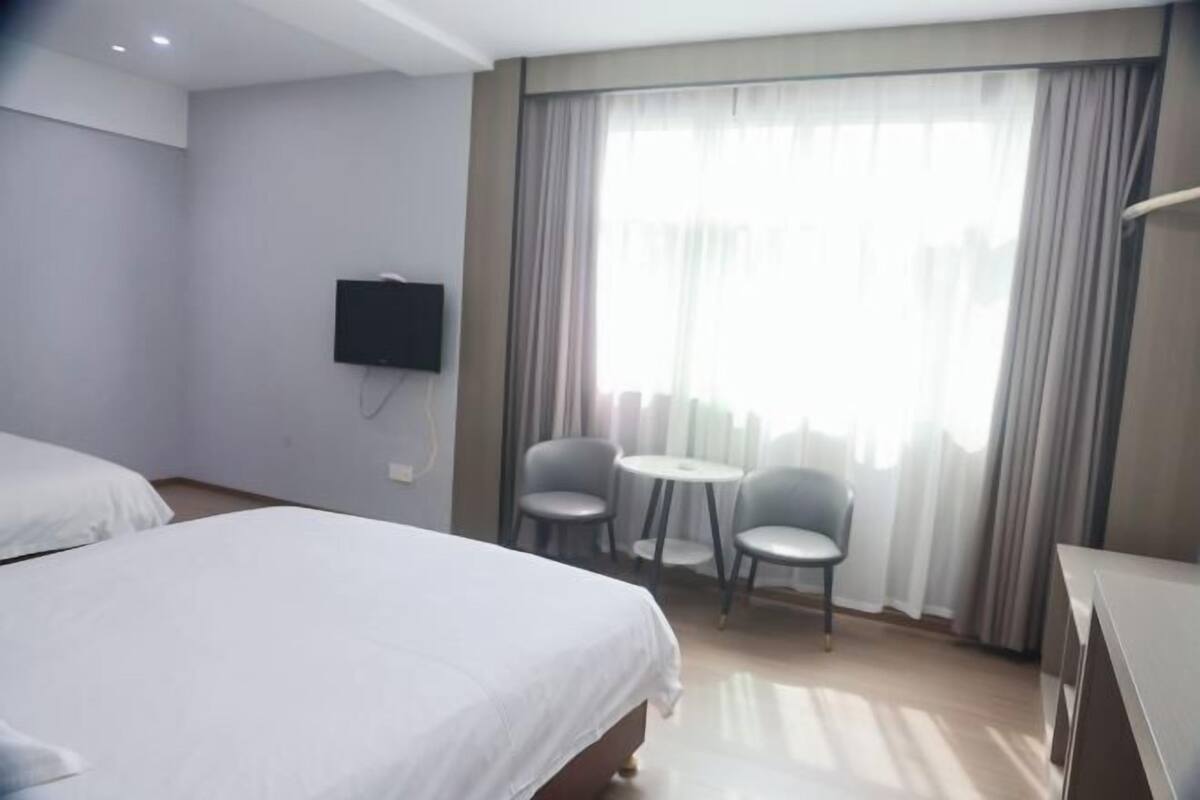 Ein Hotelzimmer mit Bett, Wandfernseher, einem kleinen Tisch mit zwei Stühlen und einem großen Fenster mit Vorhängen.