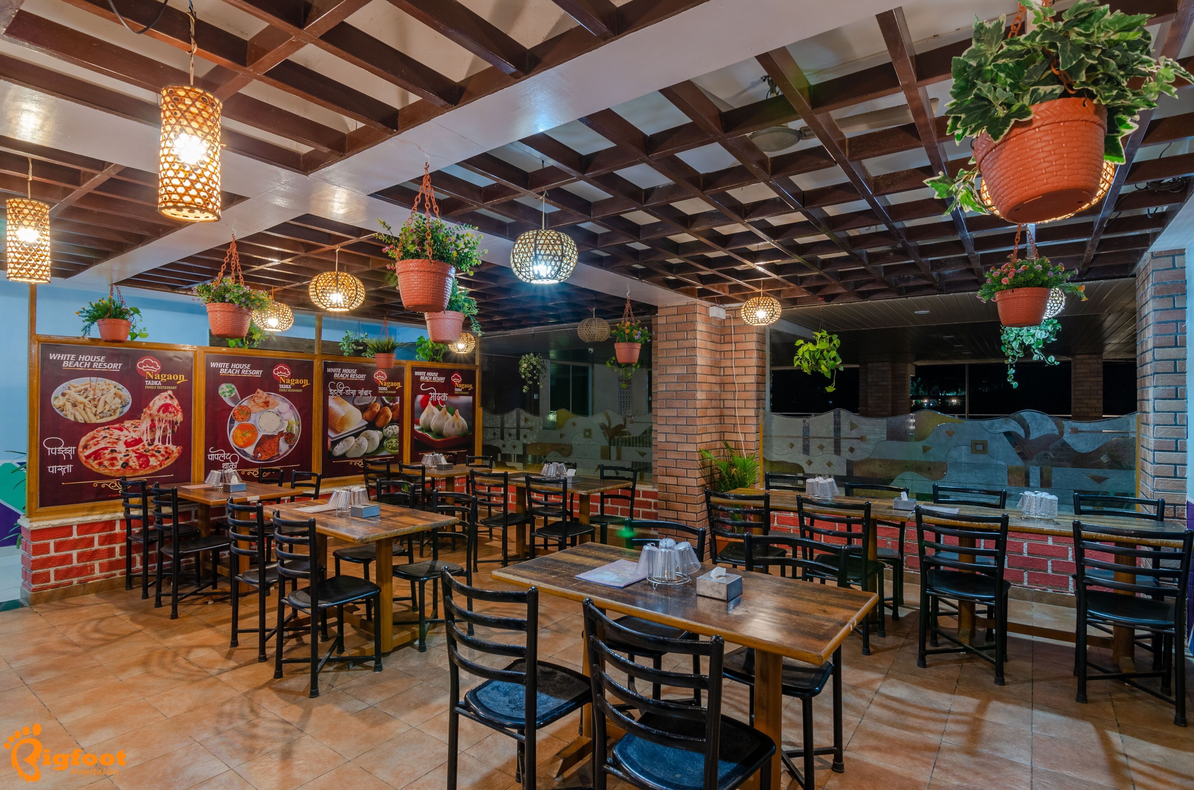 A restaurant with wooden tables and chairs, hanging plants, and a brick wall with menu boards.