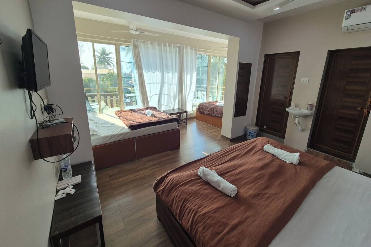 Habitación de hotel con dos camas, un ventanal grande con cortinas, una mesita de noche, un televisor montado en la pared y un ventilador de techo.
