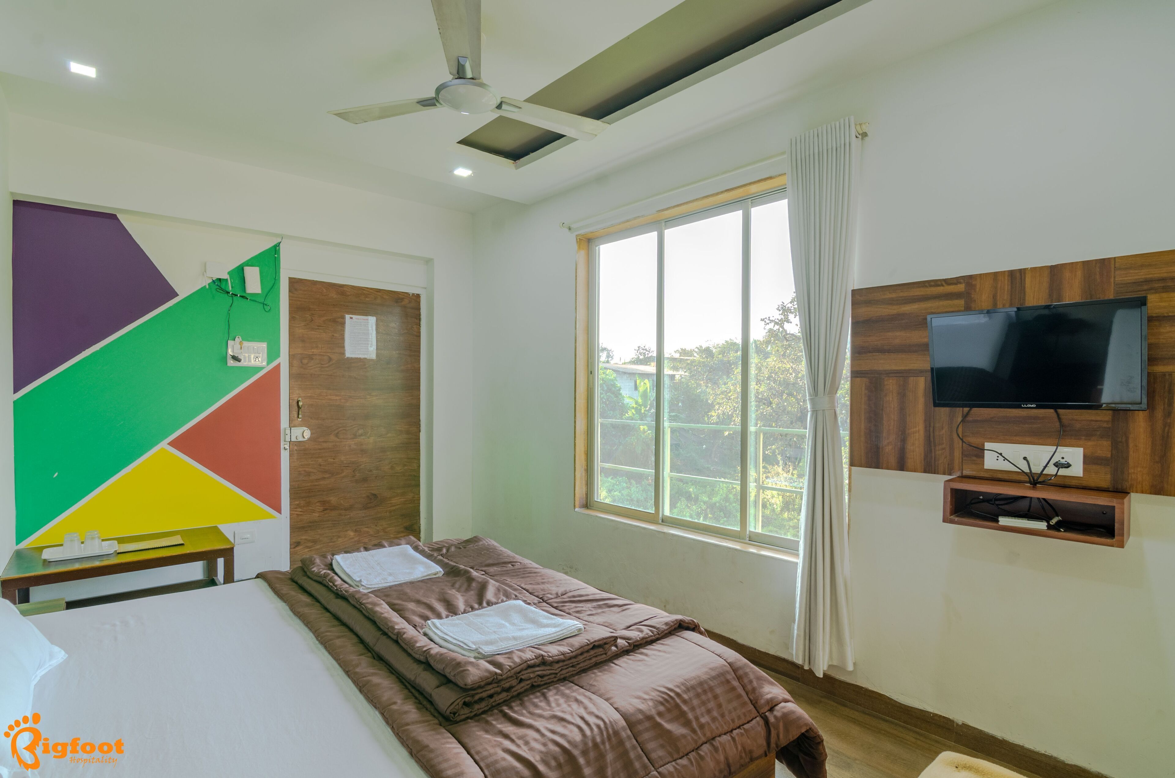 A room with a bed, a TV mounted on the wall, a window with curtains, and a ceiling fan.