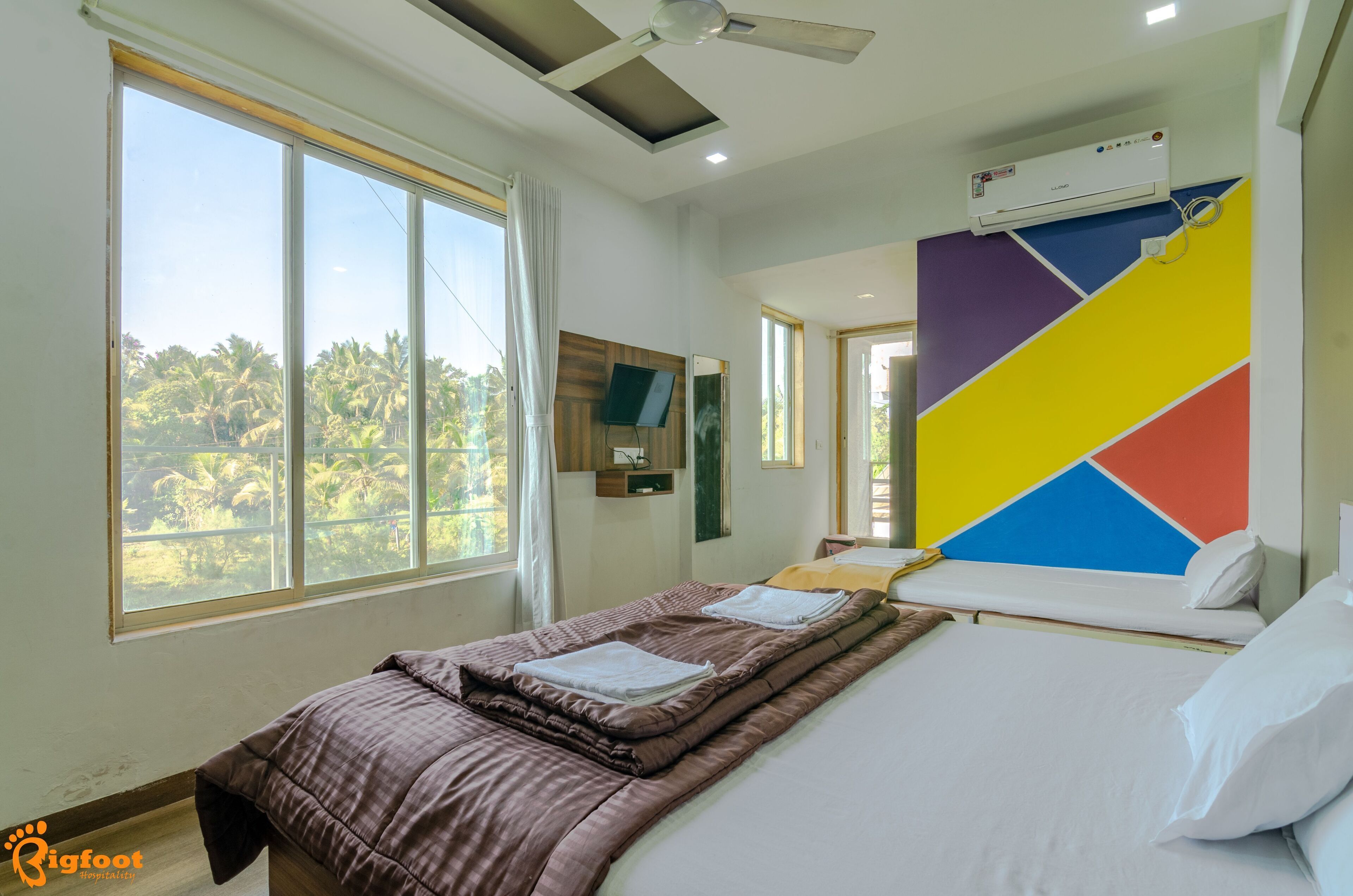 A hotel room with a bed, a TV, a window with a view of palm trees, and a colorful wall.