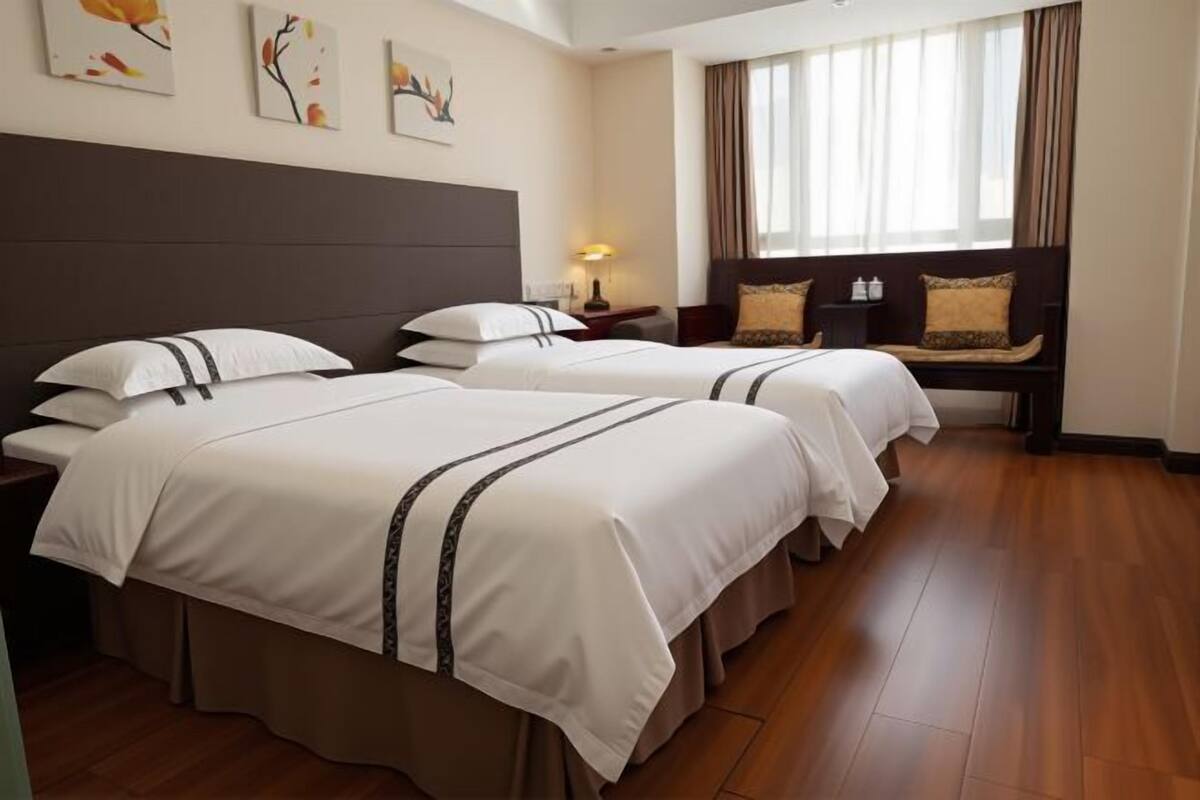 Habitación de hotel con dos camas, una mesita de noche, una lámpara, un banco con cojines y una obra de arte en la pared.