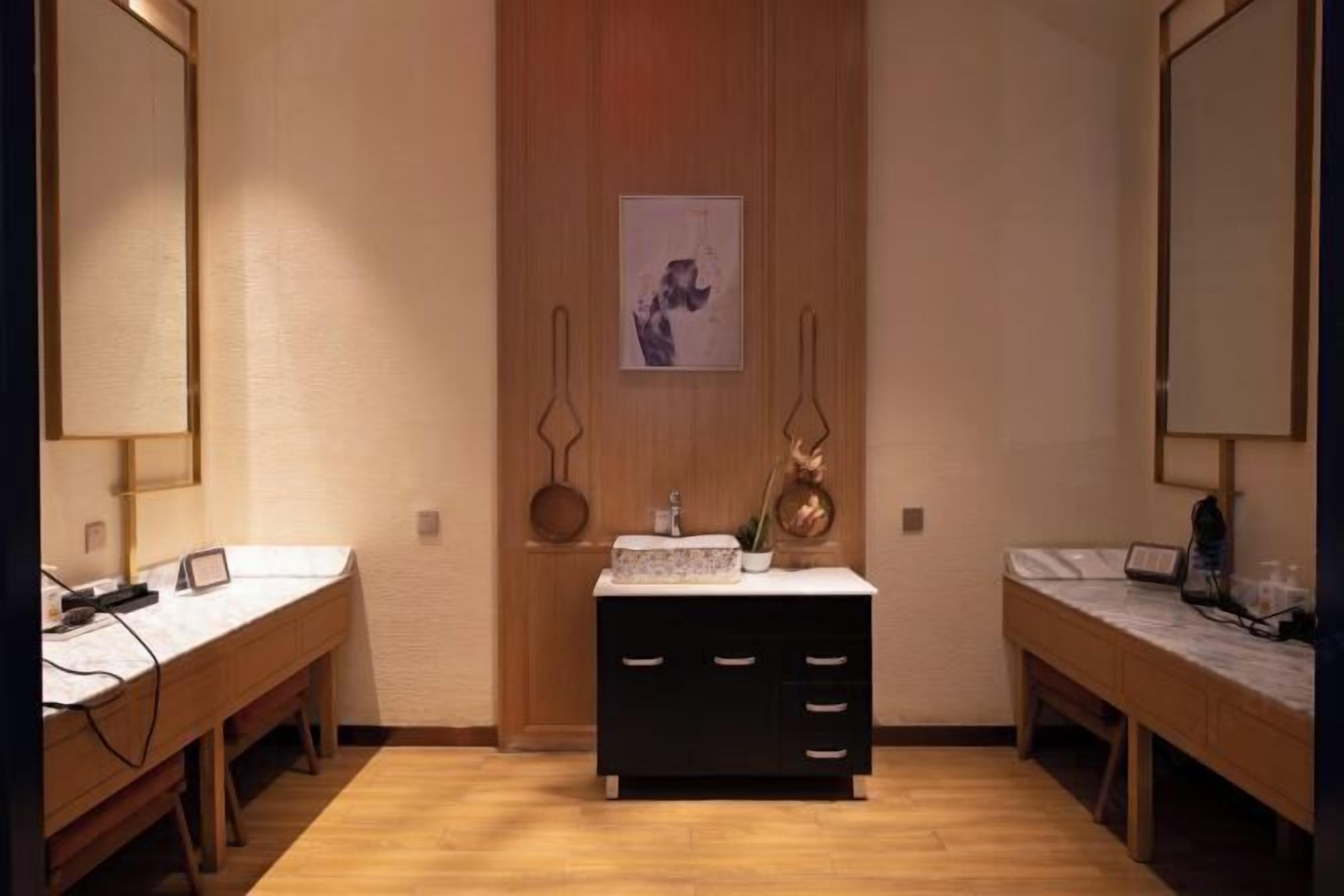 A hotel room with two wooden desks, a central black dresser with drawers, a small sink, and a framed picture on the wall.